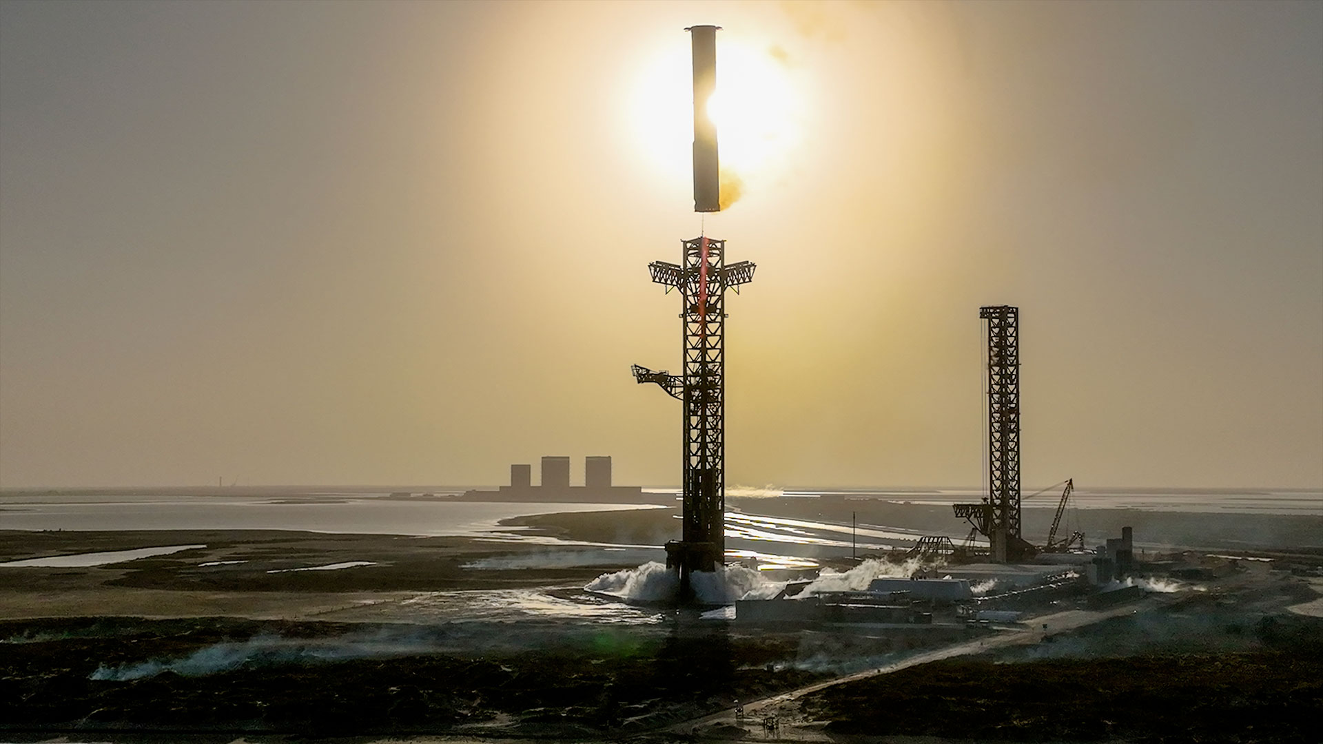 Ein Raketenbooster nähert sich seinem Startturm, um aufgefangen zu werden, während beide von der Sonne als Silhouetten abgebildet werden.