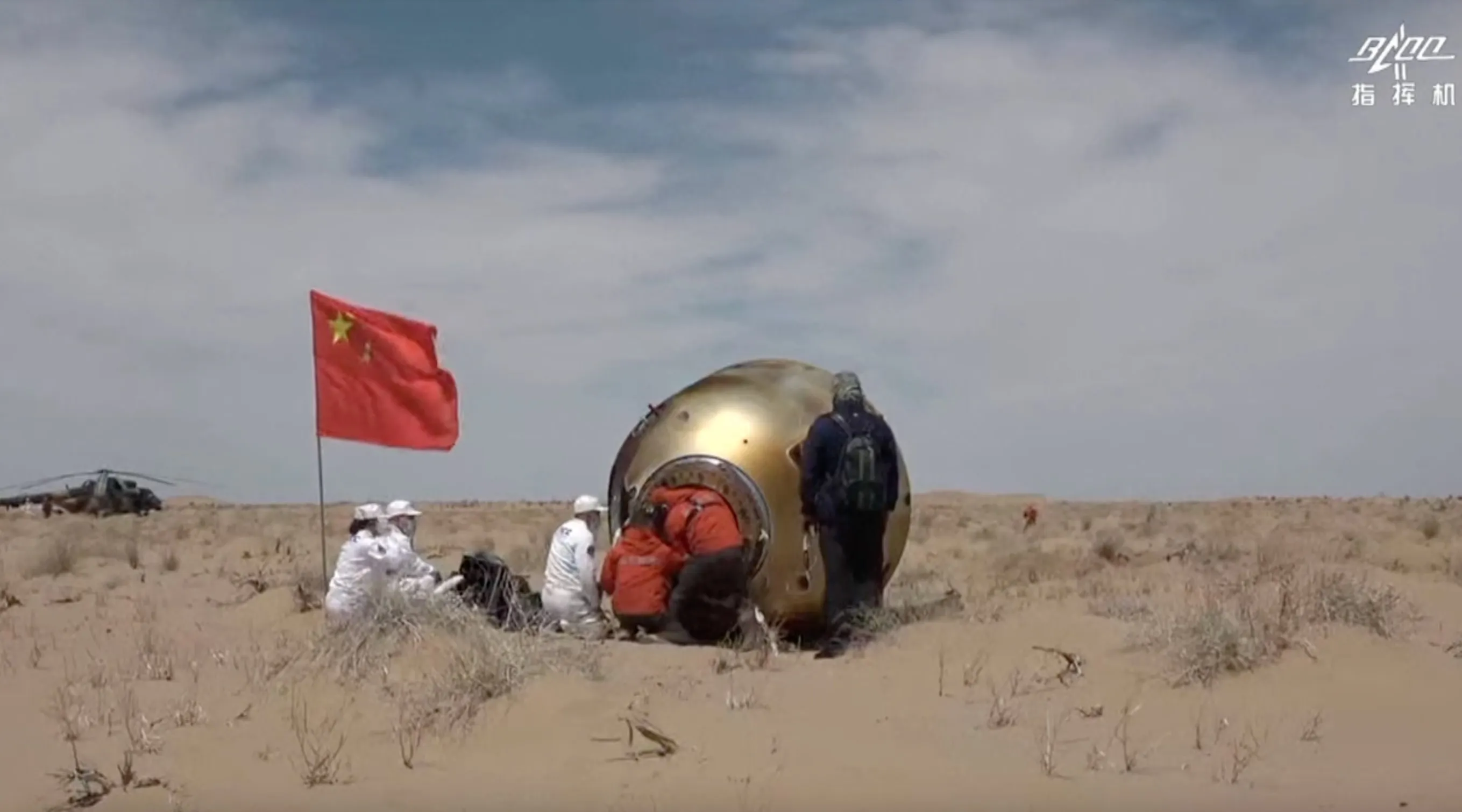 eine handvoll menschen umgibt eine goldene raumkapsel, die in einer wüstenlandschaft auf der seite liegt, in der nähe weht eine rote chinesische flagge